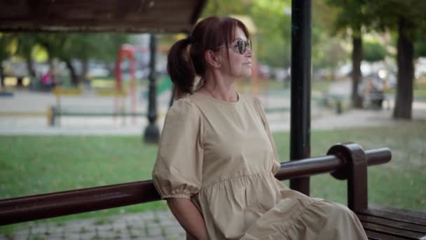 都市公園のベンチに座って話を離れて手を振っメガネで赤毛の成熟した女性 積極的な笑顔白人女性は屋外でレジャーを楽しむ — ストック動画