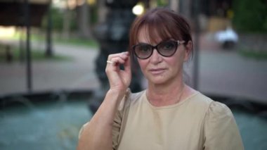 Şehir parkındaki fıskiyede poz veren kızıl saçlı emekli kadın portresi. Gözlüklü, kendinden emin beyaz bir kadın dışarıda kameraya bakıyor.