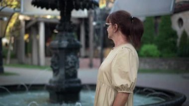 Yetişkin bir kadın kameraya bakarken kamerayı yakınlaştırıyor. Şehir parkında fıskiyenin önünde poz veren zarif beyaz kadın portresi