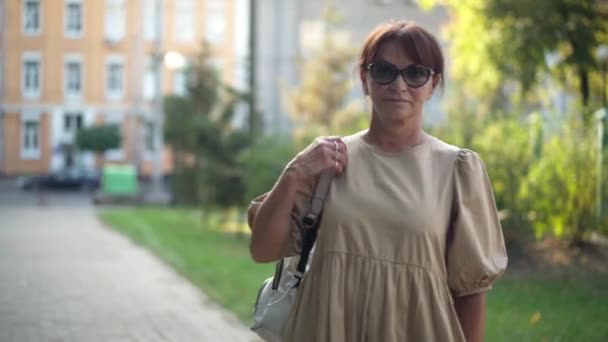 フロントビューは自信を持って成熟した女性の肩のカメラの街の公園で散歩を見てバックパックでドリーショット 女性白人退職者ポーズ屋外で歩く — ストック動画