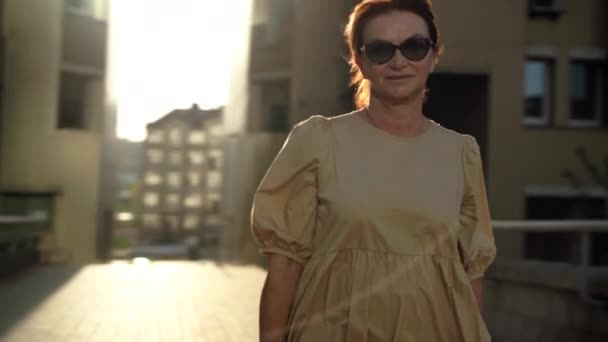 舗装道路上の都市の太陽の下で歩いて自信を持って成熟した女性の撮影パニング 白人女性の退職者屋外で夕日を楽しむ — ストック動画