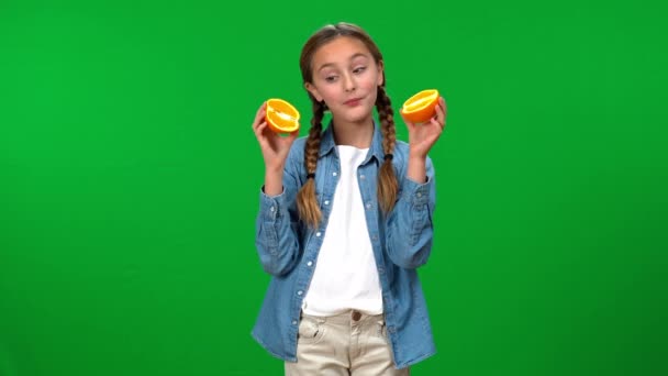 ブルネットは喜んで白人の女の子をピグテールで喜ばせ カメラを見て半分のオレンジカバーの目を置く 明るい正ティーンの肖像画ともに有機ビタミンフルーツ上の緑の画面 — ストック動画