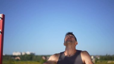 Bulutsuz mavi gökyüzünün arka planında asılı duran kırmızı jimnastik yüzükleri motive olmuş sporcu antrenmanlara başlarken. Güneşli bir günde spor yapan yakışıklı beyaz adamın portresi.