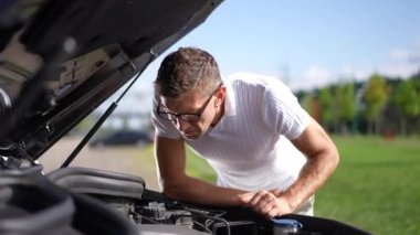 Yetişkin bir adam güneşli bir bahar gününde bozuk arabanın motorunu kontrol ediyor. Odaklanmış beyaz erkek sürücünün portresi otomobili tamir ediyor
