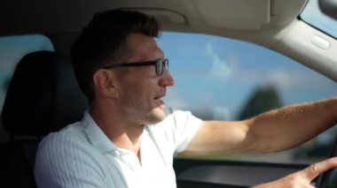Yandan bakan neşeli erkek sürücü araba kuaföründe el sallıyor, gülümsüyor ve araçtan iniyor. Açık havada güneşli bir günde otomobildeki yakışıklı beyaz adam.