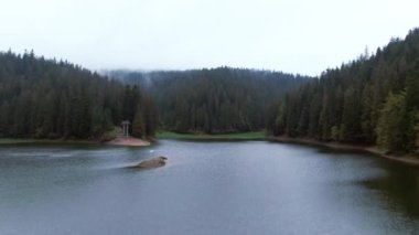 İHA, adanın ortasında Ukrayna bayrağıyla mavi Synevyr Gölü 'nün üzerine eğiliyor. Geniş açılı gölet, etrafını sisle çevreleyen yeşil kozalaklı orman.