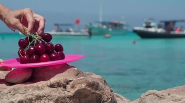 Yakın plan kadın eli, arka plandaki mavi lagünün arka planına kırmızı böğürtlen koyuyor. Tanınmayan beyaz kadın güneşli bir günde Kıbrıs tatil beldesinde vitamin atıştırmalığı hazırlıyor