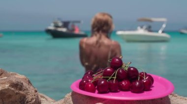 Plaj kayasında yakın plan lezzetli kırmızı böğürtlenler ve gölde yüzen sıska bir kadın yata doğru yürüyor. Kafkasyalı tanınmayan bayan turist Kıbrıs 'ta yaz tatili yapıyor