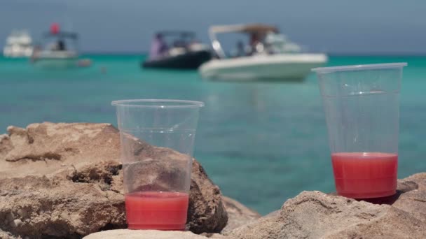 地中海沿岸の岩の上に赤いジュースを持つ2つのプラスチック製のガラスとラグーンを背景にぼやけたヨット キプロスリゾートの太陽の下でおいしい飲み物をリフレッシュ — ストック動画