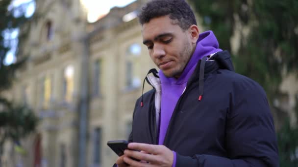 スローモーションで周りを見回すスマートフォンのメッセージングをオンラインで考えてアフリカ系アメリカ人の若者 携帯電話を屋外でハンサムな大学生の肖像画 — ストック動画