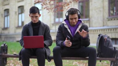 Akıllı telefon uygulama listesinden müzik dinleyen Afrikalı Amerikalı bir arkadaş olarak laptopta internette sörf yapan beyaz bir adam. Kampüste yedek kulübesinde oturan üniversite öğrencileri.