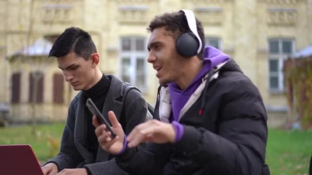 サイドアングルビューノートパソコンのキーボードに入力した白人男性は アフリカ系アメリカ人グループメイトとして ヘッドフォンのダンスベンチに座って踊る歌を集中表示します 学外の大学生 — ストック動画