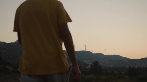 ワイドショットの夜遅い動きで歩くに入る風車と白人男性とキプロスの山々 バックビュー自信を持って男性観光客が屋外で夕暮れ散歩 — ストック動画