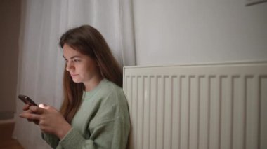 Akıllı telefon ekranında uygulamaları kaydıran ve kollarını ovuşturan güzel bir kadının yan görünüş portresi. Beyaz kadın evde soğuk radyatörde oturuyor.