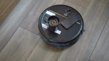 Yuvarlak süpürme robotunun yüksek açılı görüntüsü. Parke zeminde hareket eden bir bardak su ve akıllı telefon. Üst görünüm modern cihaz oturma odasını süpürüyor.