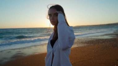 Akdeniz sahilinde altın gün batımında kameraya bakan zayıf kadının portresi. Kıbrıs 'ta okyanus kıyısında güneş ışığında poz veren, kendinden emin Kafkasyalı turist