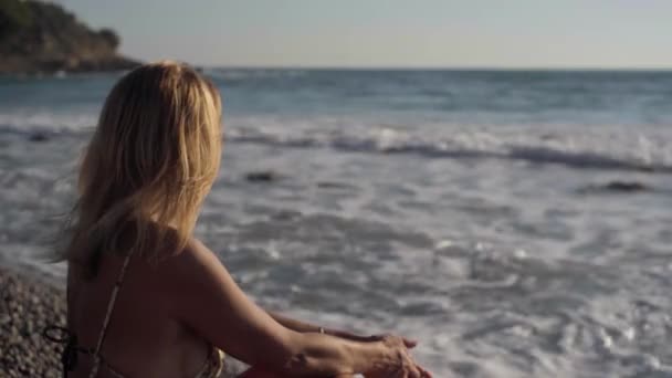 ブロンドの日焼けした女性は地中海の波が海岸を転がるようにビーチの左側に座っています キプロスで休暇を楽しむリラックスしたコーカサス人気楽な観光客 — ストック動画