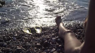 Sıska kadın bacağı sağda ve günbatımı güneşi arka planda Akdeniz dalgalarını yansıtıyor. Tanınmayan Kafkasyalı turist, Kıbrıs tatil beldesinde gün batımının keyfini çıkarıyor