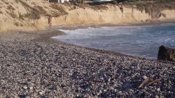 地中海的卵石海滩 背景是戴着太阳镜和比基尼的晒黑了的模糊女人 在塞浦路斯平静的阳光明媚的海湾 海浪在海岸上翻滚 游客们在户外愉快地漫步 — 图库视频影像