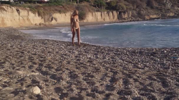 広いショットスリム日焼けした白人ブロンドの女性が地中海の小石のビーチを散歩離れる 晴れた日にキプロスリゾートの太陽の下で幸せなリラックスした観光客の散歩を自信を持って — ストック動画