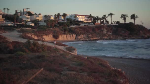 地中海の波が黄金の夕日で海岸に転がり 白人女性がビーチに立ってカメラを見て手を振っています 観光客の呼び出しと夕暮れの美しいキプロスリゾートを撮影 — ストック動画