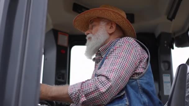 蓄着胡子的老人坐在拖拉机小木屋里转动方向盘 集中的严重农民驾驶农业收割机到户外 — 图库视频影像