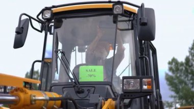 Son sınıf öğrencisi traktör kabininde% 70 indirim duyurusuyla oturuyor. Beyaz erkek çiftçi yeni ekiciyi yavaş çekimde seçiyor.