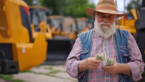 满意地微笑着的老人站在室外数钱 背后有黄色的拖拉机 快乐的白人卖家出售农业机械赚取收入的肖像 — 图库视频影像