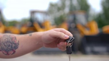 Yakın plan tanınmayan dövmeli genç adam sarı ekicilerin arka planında kıdemli alıcıya anahtar veriyor. Kafkasyalı satıcı ve alıcı araç anahtarını açık havada ağır çekimde değiş tokuş ediyorlar.