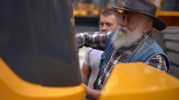 老人打开前部的巨大拖拉机与年轻人在室外交谈 有经验的专业白人农民和同事一起选择新车 慢动作 — 图库视频影像