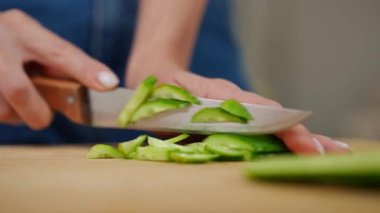 Yakın plan kadın eli, yeşil salatalığı bıçakla kesiyor. Yakından tanınmayan genç beyaz kadın mutfaktaki sağlıklı sebzeleri kesiyor.