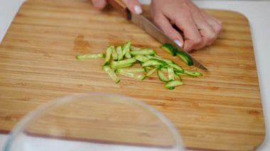 Yakın çekimde tanınmayan bir kadın salatalığı bıçakla kesiyor ve cam şeffaf kaseye salata malzemesi koyuyor. Genç, zayıf, beyaz bir ev hanımı evde lezzetli yemekler pişiriyor.