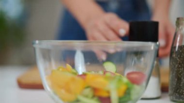 Salatayla yakın çekim kasesi ve yavaş çekimde beyaz tuz serpiştiren tanınmayan genç bir kadın. Kapalı, sağlıklı, lezzetli akşam yemeği mutfaktaki masada.