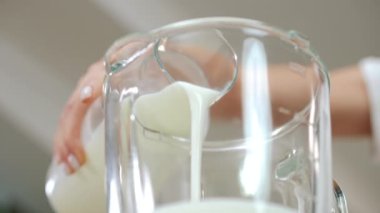 Kadın eli blender 'a beyaz süt dökerken yavaş çekimde ayrılıyor. Yakından tanınmayan genç bir kadın sabahları evde sağlıklı meyve suyu karıştıran yoğurt ekliyor.