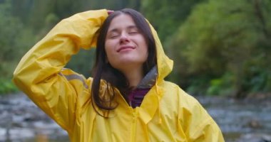 Ön manzara, gülümseyen mutlu genç kadın yağmurluğunu çıkarıyor ve ağır çekimde yukarı bakıyor. Dışarıdaki dağ ormanlarında tasasız beyaz turist portresi. Sinema 4k ProRes Merkezi