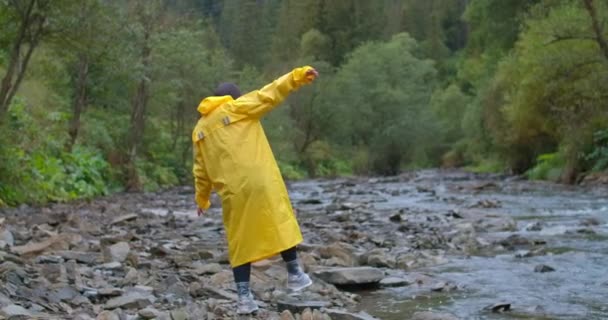 バック遅い動きで山の川の岩の上を歩く黄色のレインコートで女性観光客を表示します 広いショット自信を持って白人の若い女性の森の中で石の上を歩く シネマ4K Prores — ストック動画