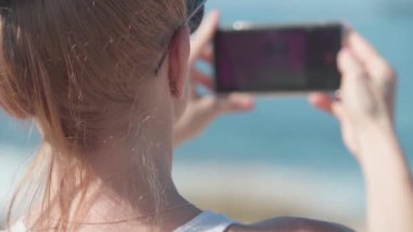 Akdeniz dalgalarını çeken sarışın bir turistin omzunun üstünden akıllı telefon uygulaması gün ışığında duruyor. Arka plan. Tanımlanamayan beyaz kadın yaz tatilinin tadını çıkarıyor.