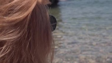 Güneş gözlüklü kızıl saçlı beyaz kadının yakın plan fotoğrafı. Arka planda berrak körfez suyu var. Kıbrıs 'taki Akdeniz plajında kendinden emin mutlu turist