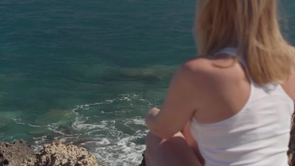 Kristalhelder Oceaanwater Spatten Rotsachtige Kust Met Wazig Blonde Vrouw Zitten — Stockvideo