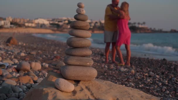 地中海のビーチにある石のクローズアップ塔と 背景に立っている大人のカップルを愛しています 幸せなリラックスした白人男性と女性を受け入れます楽しいです黄金の夕日オンキプロス — ストック動画