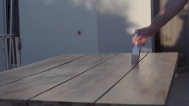 Ahşap masa üstü ve erkek eli sağda fırçayla boya sürüyor. Yakından tanınmayan beyaz bir adam ahşap masayı boyuyor.