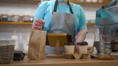 Kahve fincanlarını yakın plan çek ve kafeteryadaki beyaz kadın ellerinde kağıt poşet getir. Kahve dükkanında içki ve tatlı içen tanınmamış genç barista.