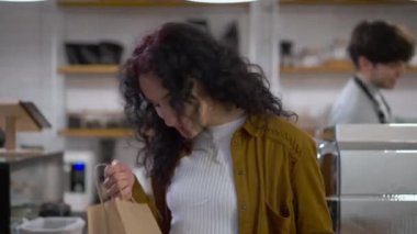 Yemek siparişi veren mutlu beyaz kadın kameraya bakıp arka plandan geçen garson gibi gülümsüyor. İçerideki kafede poz veren kendinden emin müşterinin portresi