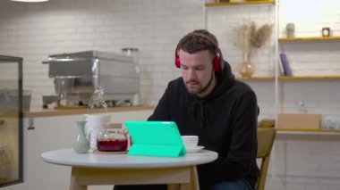 Kulaklıklı genç adam, kafeden geçen kadınlar olarak dijital tabletten çevrimiçi video izlerken kahve içiyor. Şehir merkezinde uzaktan çalışan konsantre beyaz erkek serbest çalışanın portresi.