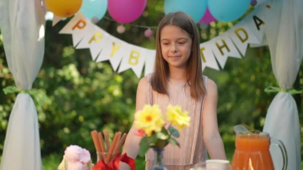 笑容满面的白种人女孩站在公园的餐桌旁 背靠着用气球装饰的生日快乐 公园里心满意足的孩子们的正面画像 — 图库视频影像