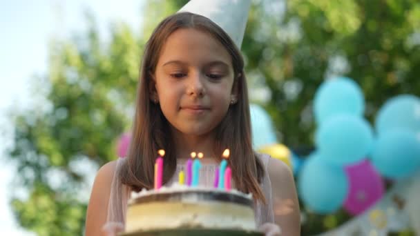 在生日蛋糕上吹灭蜡烛的快乐女孩的特写 动作缓慢 面带微笑 在阳光明媚的夏日 白人孩子们在公园外面庆祝节日 他们对此感到非常满意 — 图库视频影像