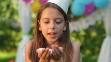 Neşeli mutlu kız yavaş çekimde konfeti üflüyor ve kameraya yakından bakıyor. Güneşli bir yaz gününde, doğum gününde dışarıda poz veren neşeli, beyaz bir çocuğun yakın plan portresi.