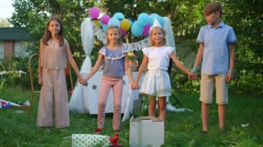 Ağır çekimde zıplayan neşeli çocuklar el ele tutuşup gülümseyen kameralara bakıyorlar. Neşeli beyaz erkek ve kızlar yazın arka bahçesinde doğum günü partisi veriyor.