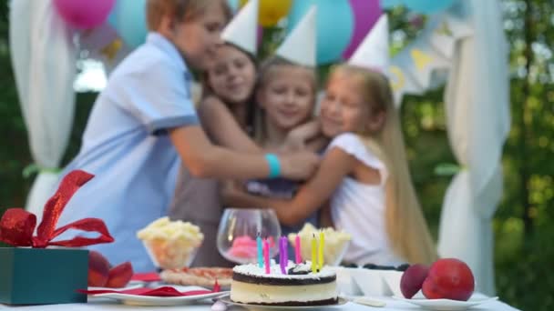 背景の遅い動きに抱きつくぼやけた子供たちのグループと裏庭のテーブルの上の誕生日ケーキ 屋外でお祝いを受け入れる幸せな子供たちとのクローズアップおいしい甘いデザート — ストック動画