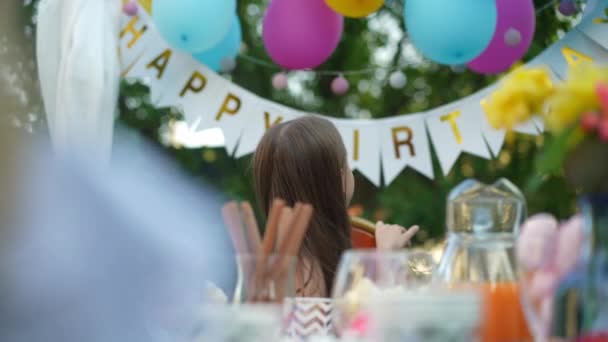 快乐的女孩回头看派对装饰坐在户外的生日桌旁 一个快乐 快乐的高加索小孩在夏天的后院慢吞吞地四下张望的画像 — 图库视频影像